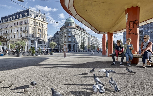 Zentralplatz in Biel: Im Hintergrund fassen Gebäudefassaden den Platz ein und es gibt ein Restaurant mit Aussenbestuhlung. Im Vordergrund sitzen eine Frau und zwei Kinder in einer Tramhaltestelle mit Bank, roten Stützen und weitläufiger Überdachung. Sie füttern Tauben. 